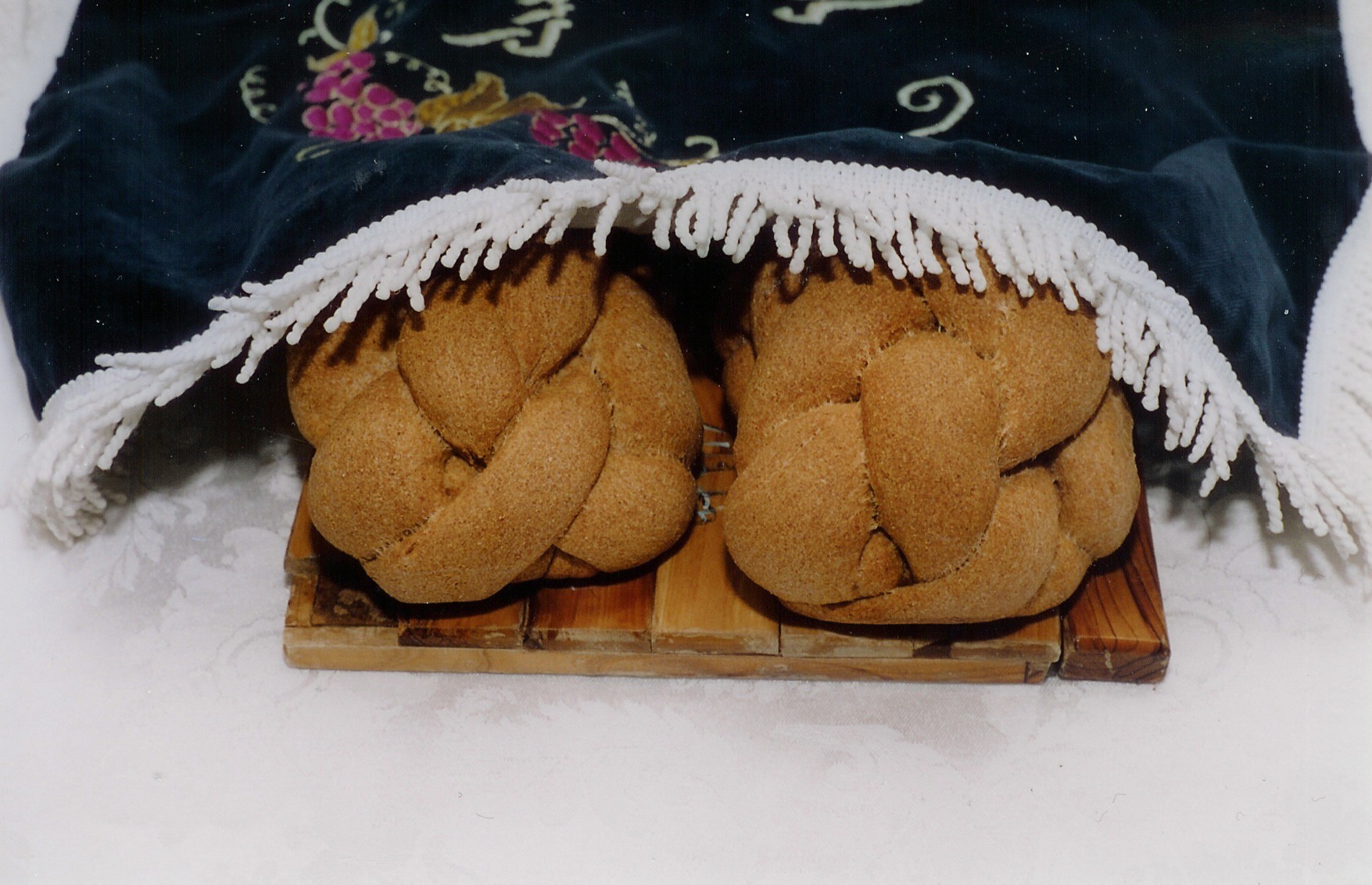 Two braided challah loaves on a wooden board under an embroidered cover