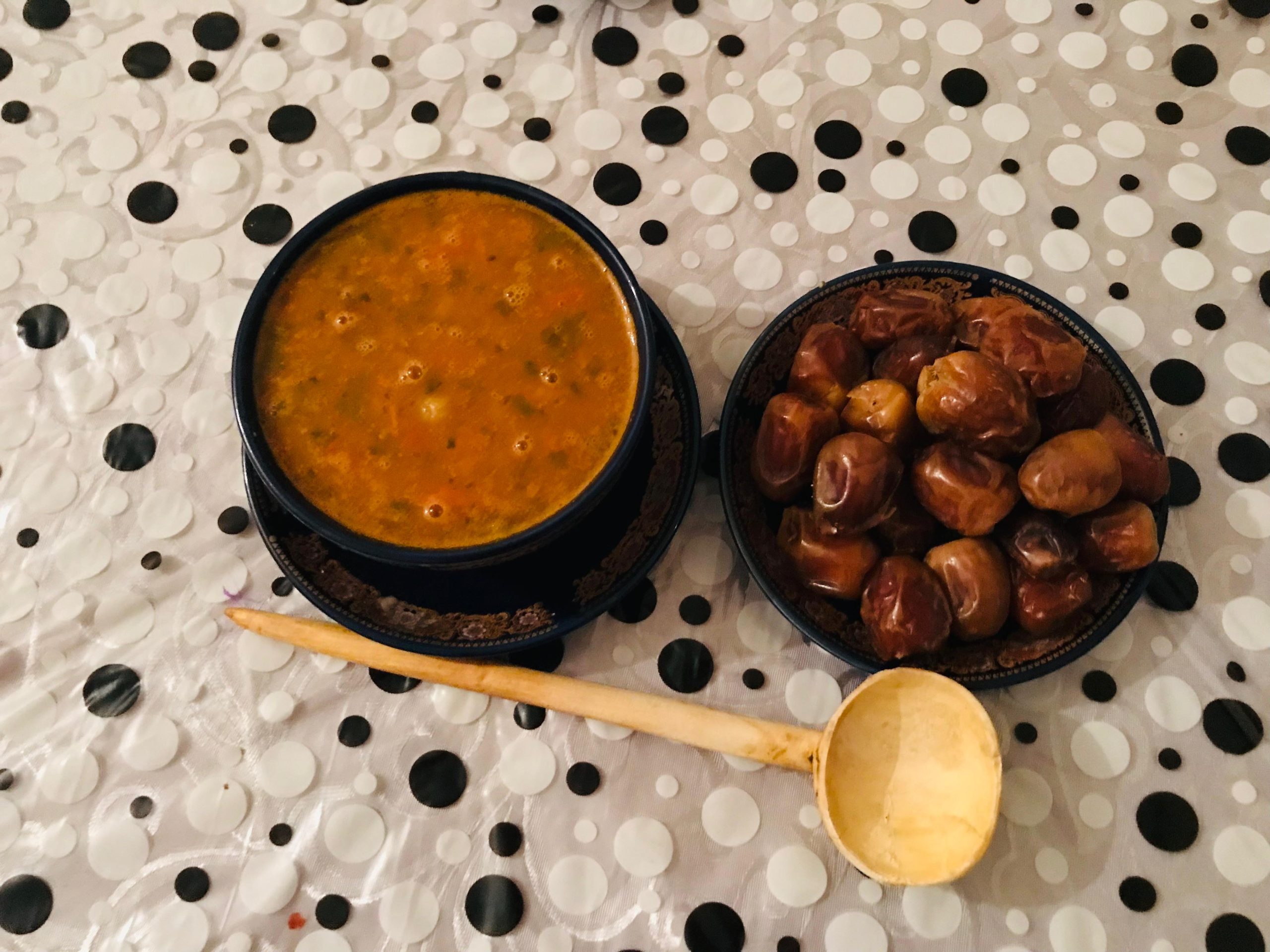 A bowl of harira soup beside a dish of dates and a wooden spoon on a patterned tablecloth