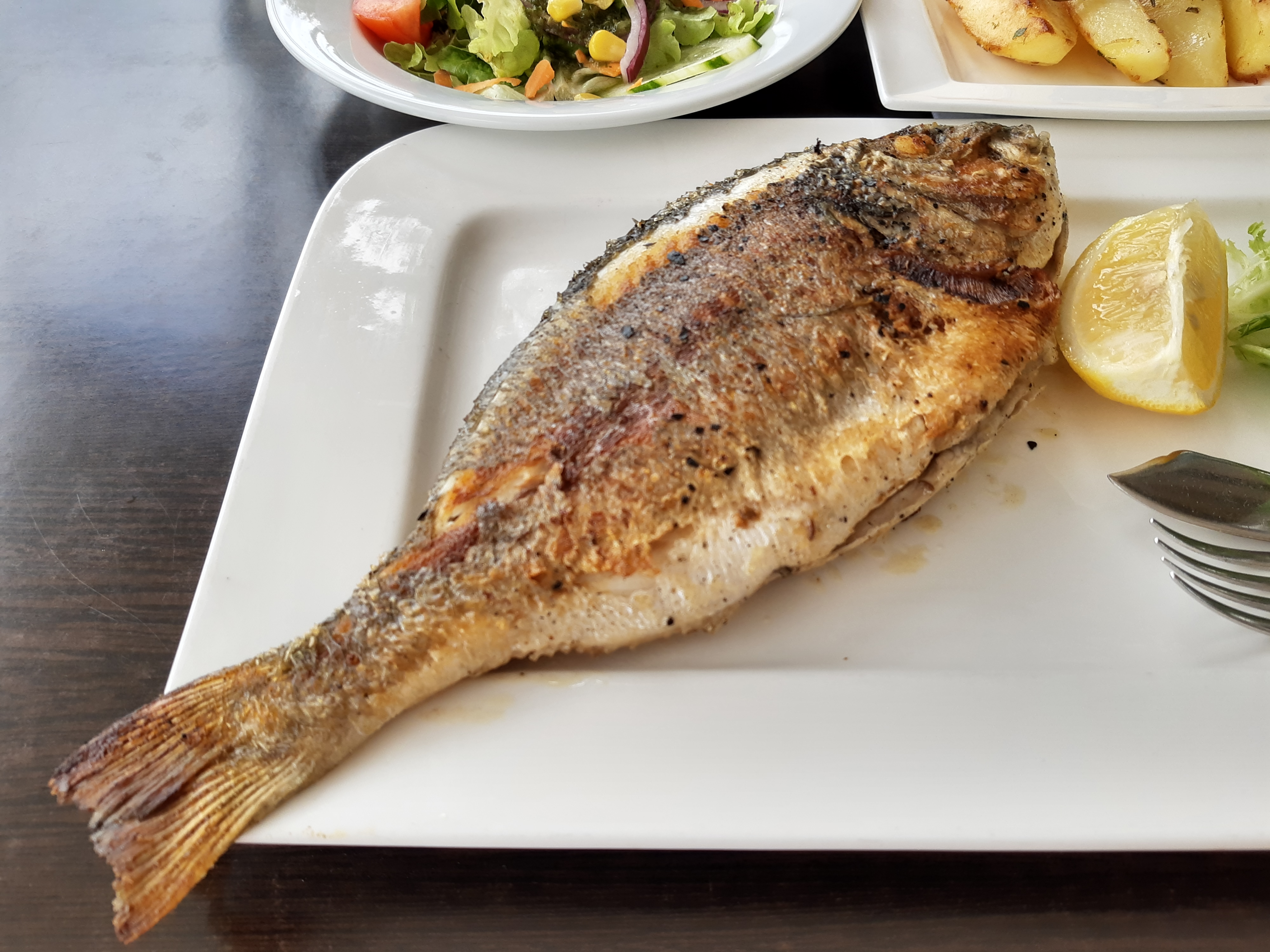 A whole grilled sea bream with a lemon wedge, with a side salad and potatoes behind