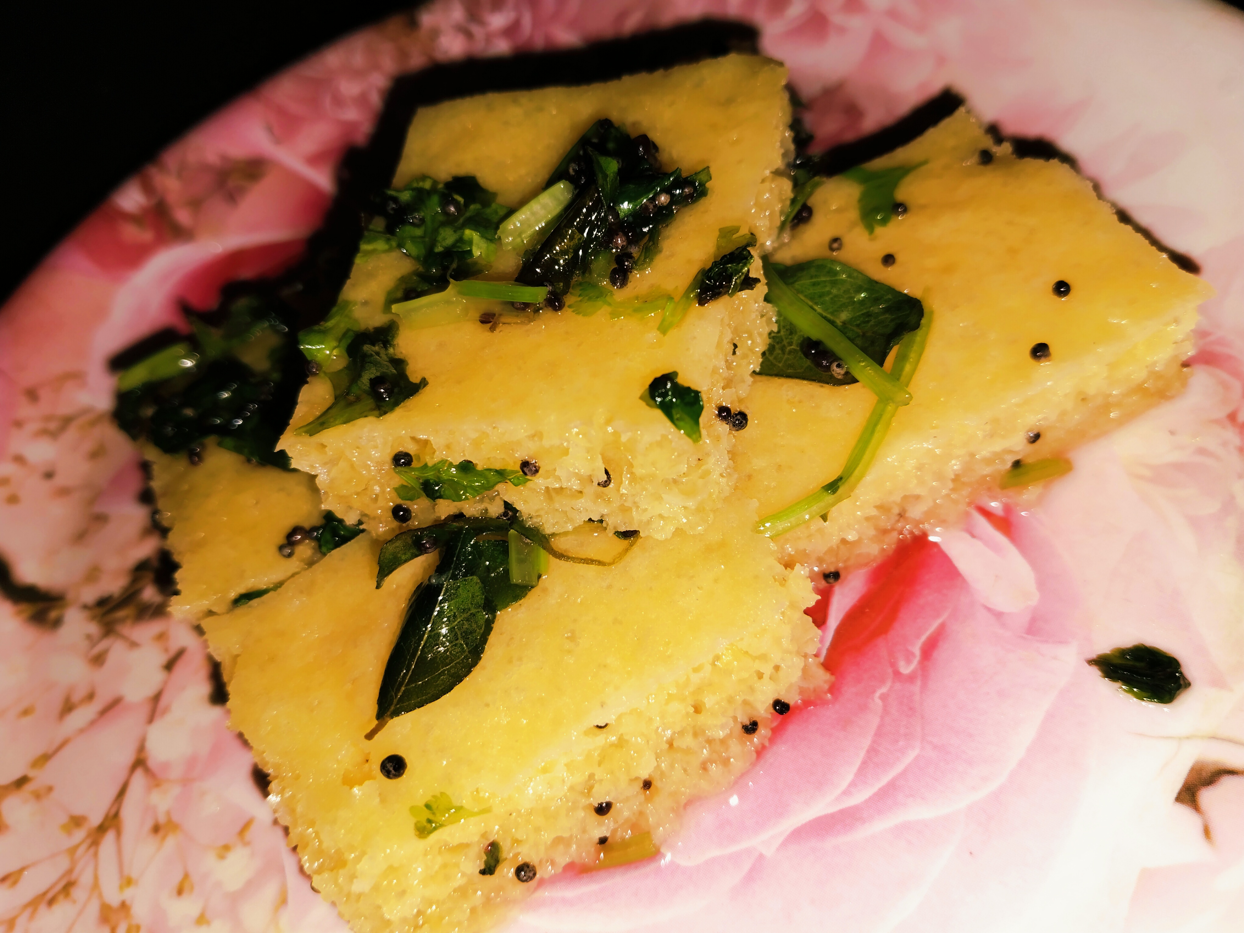 Squares of steamed dhokla topped with mustard seeds and curry leaves on a floral plate