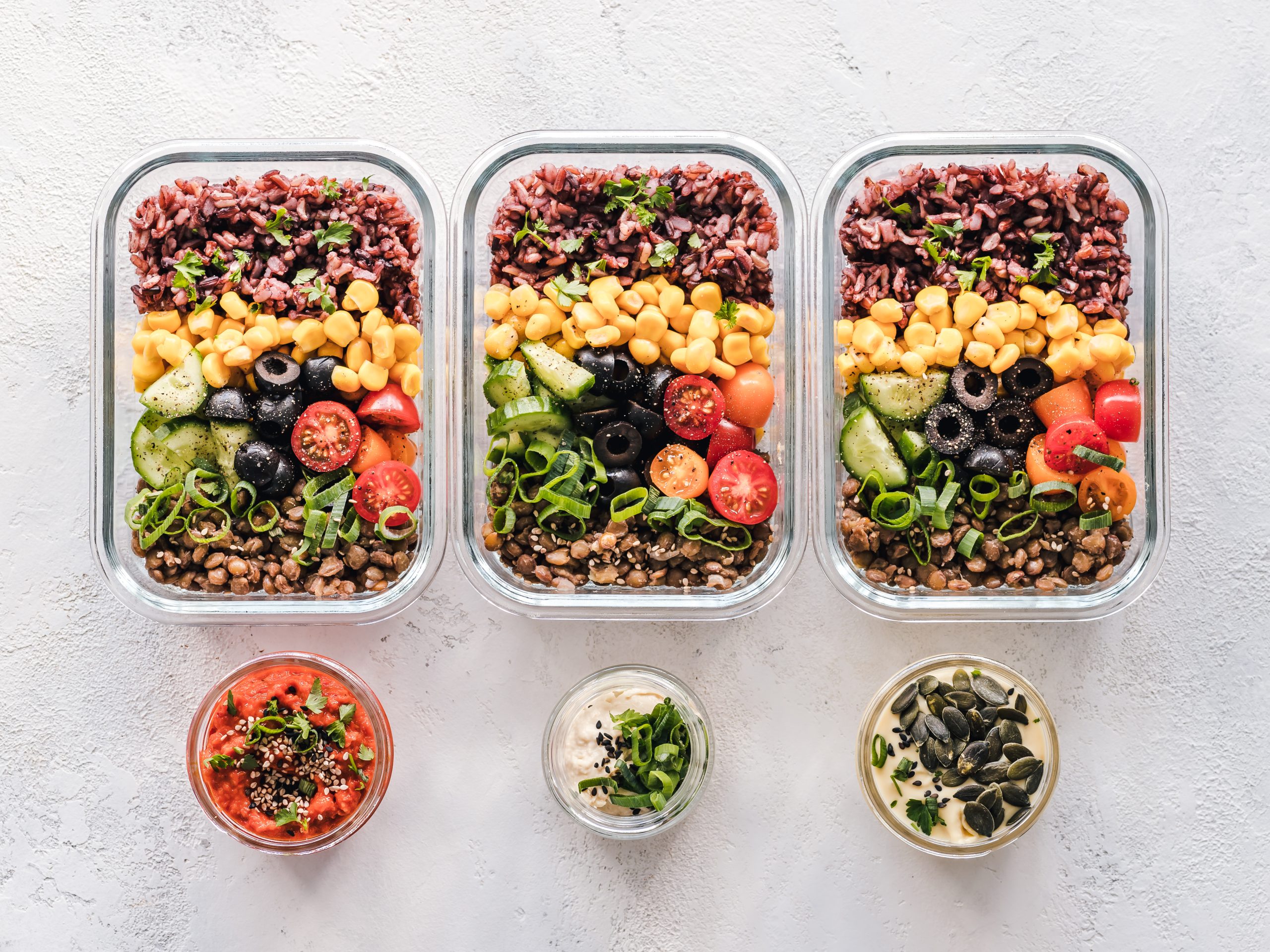 Three glass meal prep containers of rice, lentils, sweetcorn, tomato and cucumber with small pots of dips