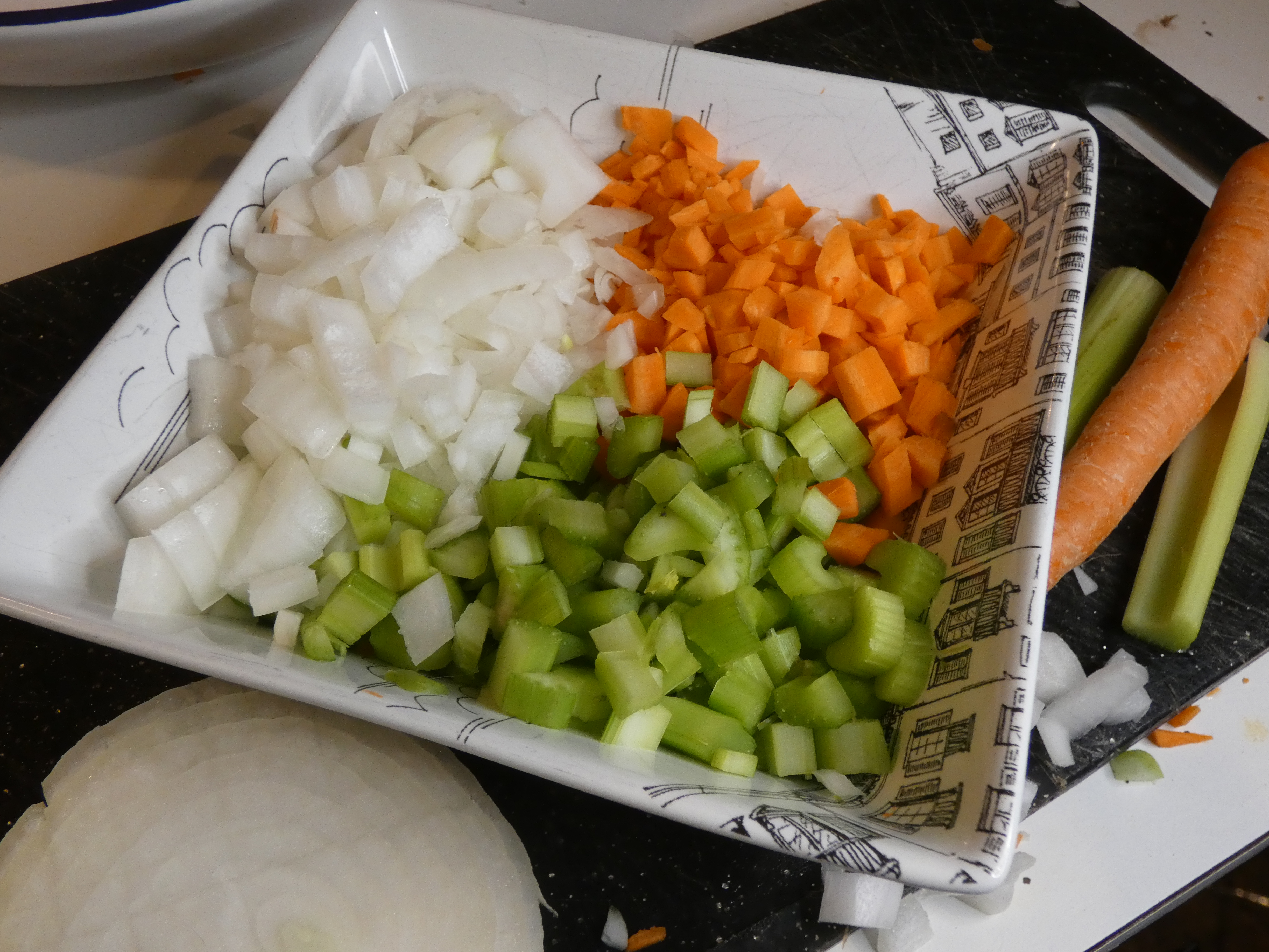 Diced onion, carrot and celery arranged on a square serving plate beside a cutting board