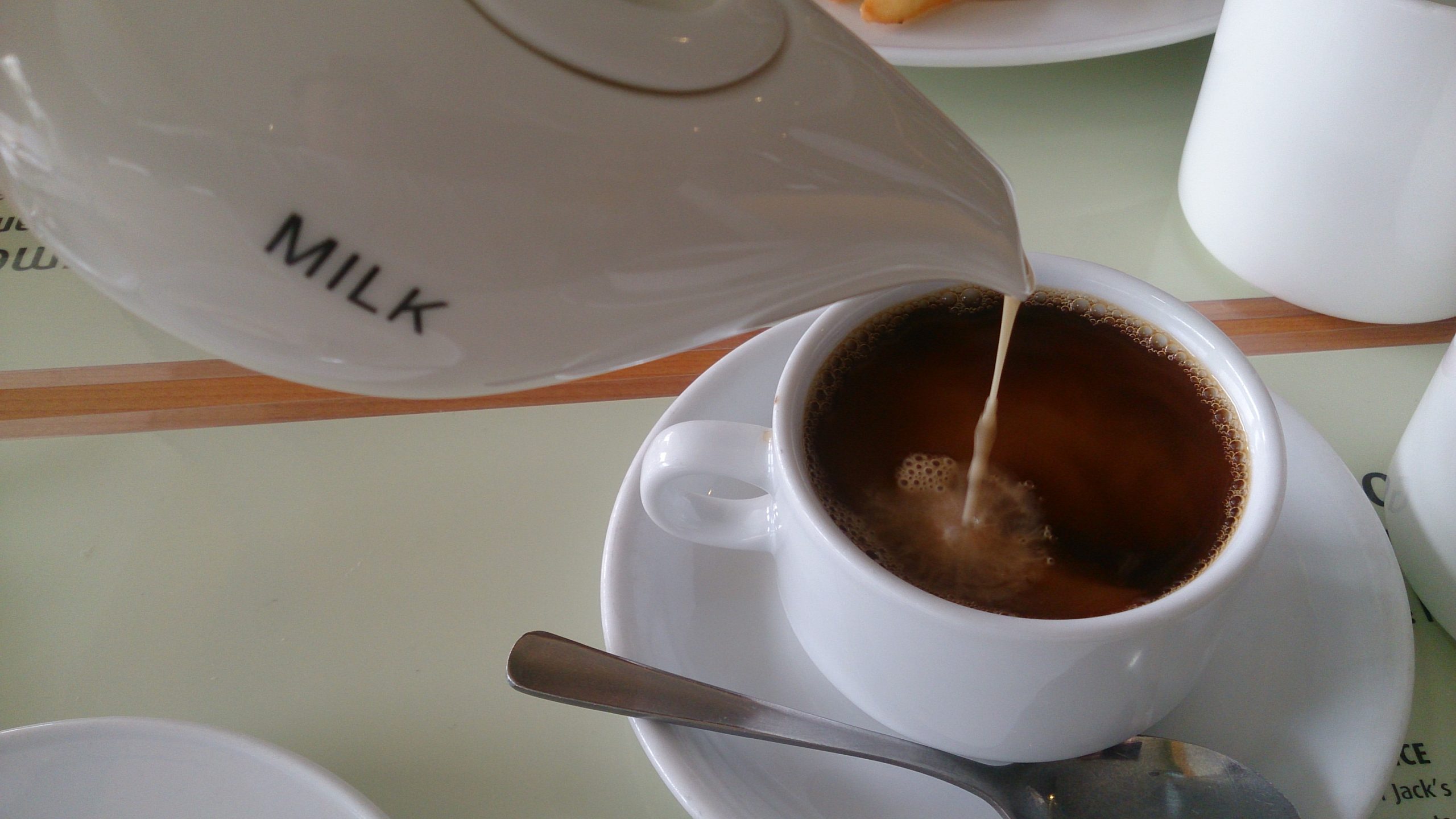 Milk being poured from a small jug into a cup of black coffee at a cafe table