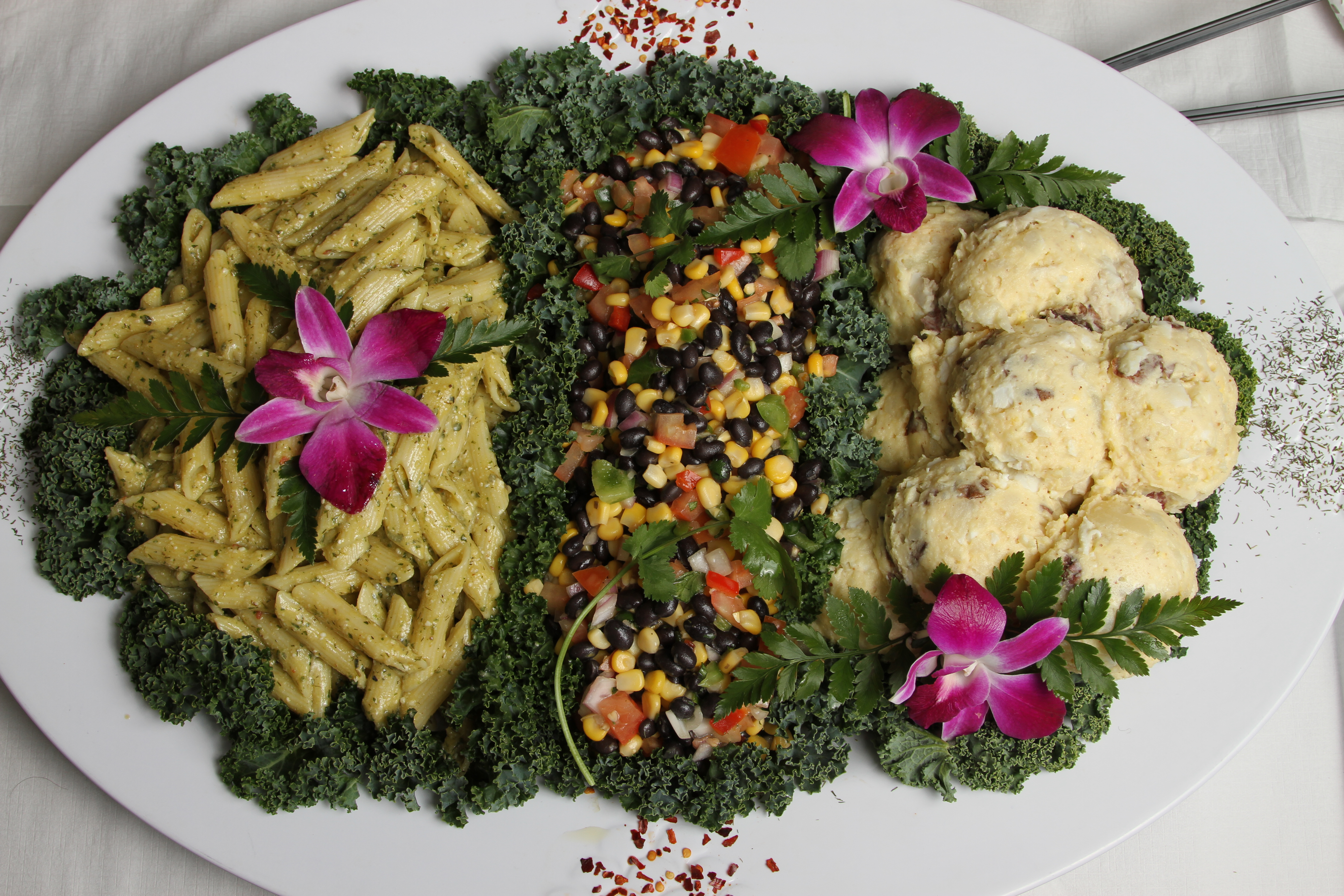 Large platter with separate sections of pesto pasta, black bean salad and potato salad on kale