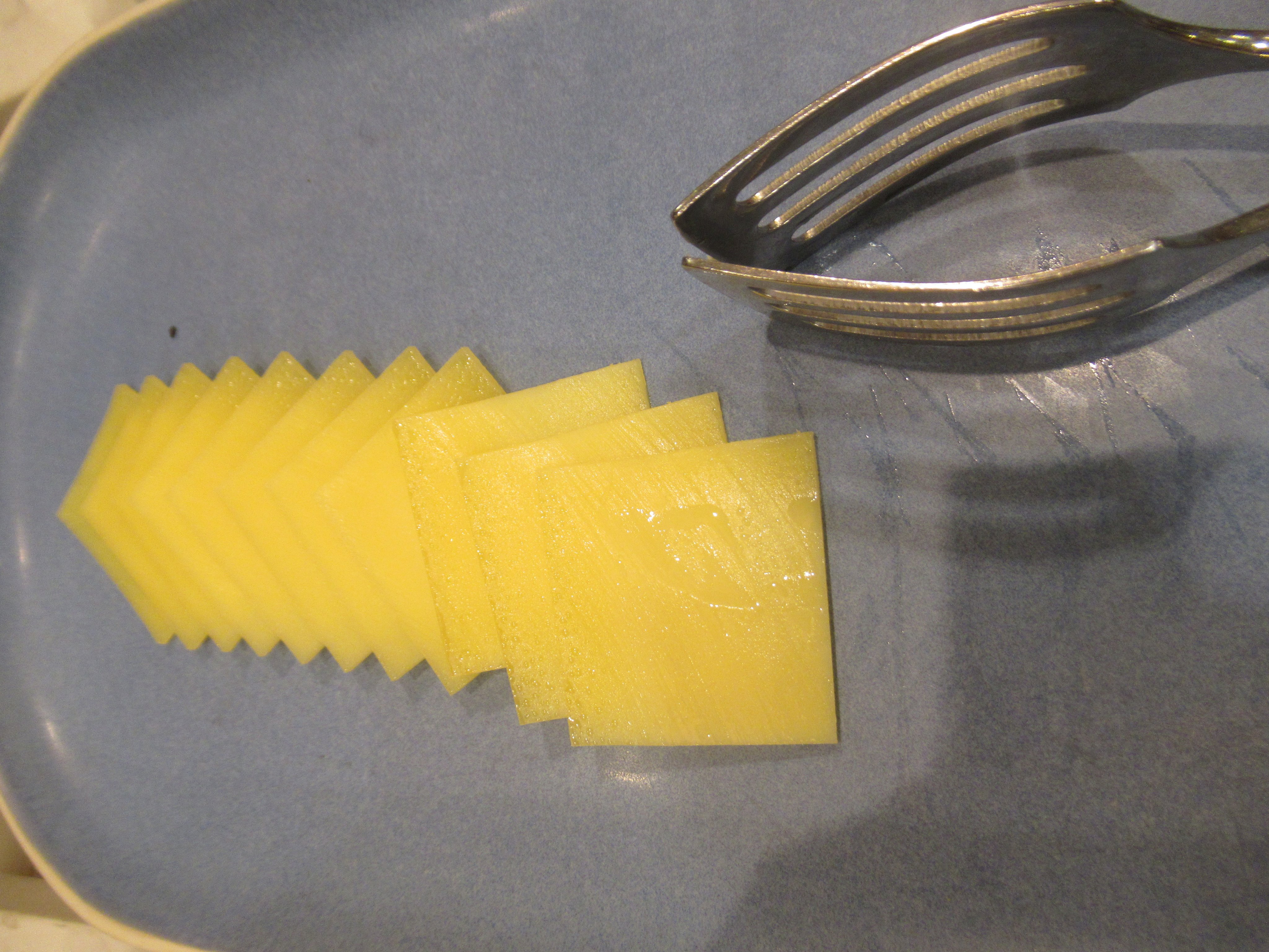 Thin squares of hard cheese fanned out on a blue plate beside serving tongs